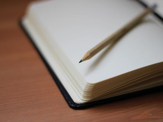 A blank notebook open with a pencil laid on a wooden desk. 