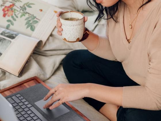 Woman with glasses holding teacup reads from a laptop